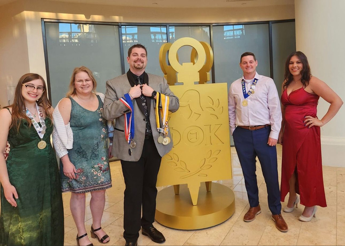 Four Phi Theta Kappa student members and one advisor pose in formal attire in front of a large gold PTK emblem at a national convention. The students wear medals and honor cords, smiling proudly. The background features glass windows and a polished marble floor.