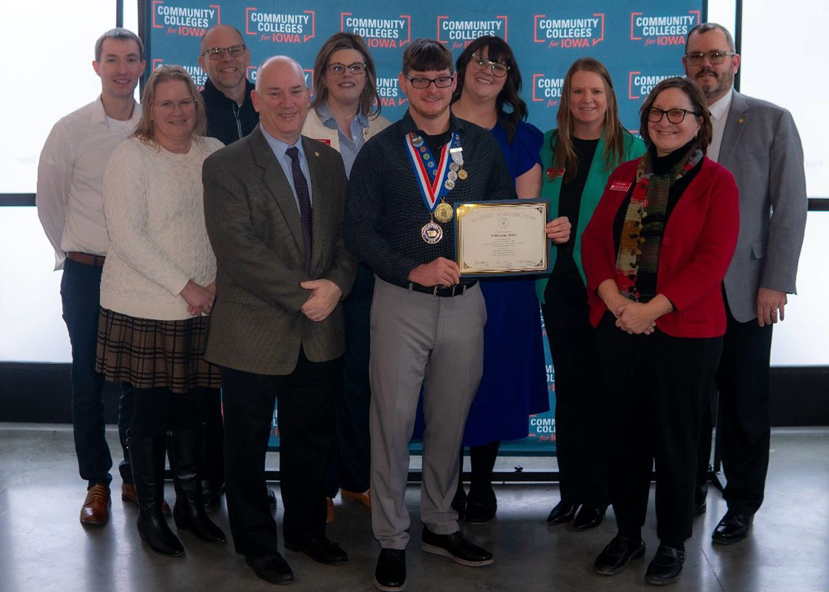 A group of college faculty, staff, and supporters pose with Christopher Bolin. Bolin stands in the center holding an All-Iowa Academic Team certificate, wearing medals and pins. Behind them is a blue banner reading “Community Colleges for Iowa.” Everyone is smiling in celebration of the recognition.