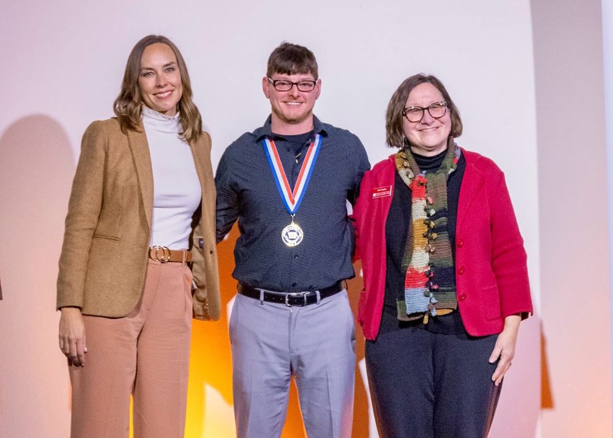 Christopher Bolin stands on stage between two women, smiling and wearing a medal around his neck at a PTK All-Iowa Academic Team awards ceremony. All three are dressed professionally and facing the camera.