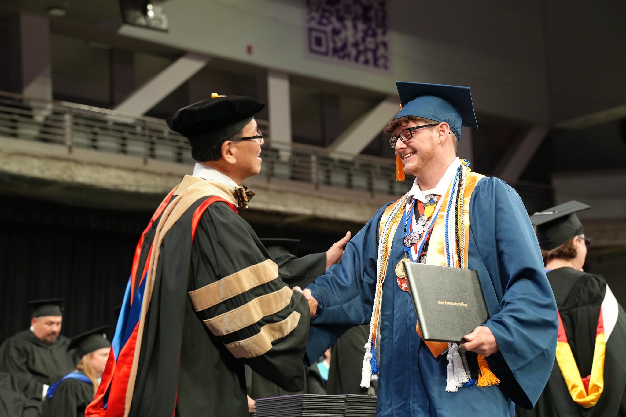 Christopher Bolin, 2025 Scott Community College graduate, smiles as he receives his diploma on stage at commencement. Dressed in his cap and gown, he wears multiple honor cords and medals while shaking hands with EICC Chancellor Dr. Liang Chee Wee.