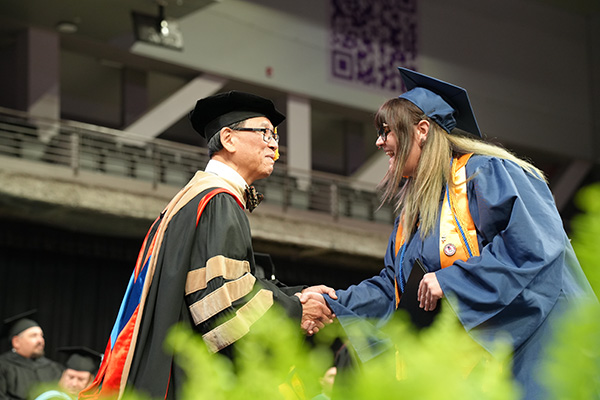 Joeanna Moore enthusiastically shakes hands with EICC Chancellor Dr. Wee in a bowtie and graduation regalia, surrounded by fellow graduates on stage.