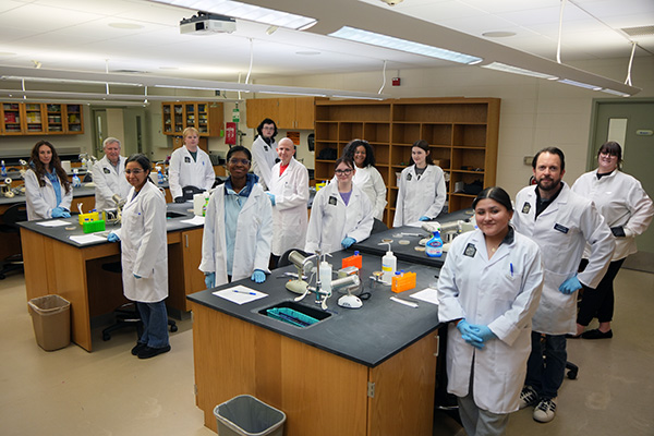 A group of students and instructors wearing white lab coats stand smiling in a science lab at SCC, surrounded by lab benches and equipment.