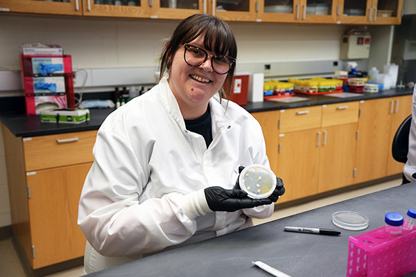 Joeanna Moore is dressed in a white lab coat and black gloves smiles and holds up a petri dish containing samples, seated at a lab table in a college science classroom.