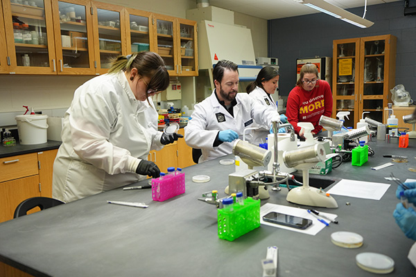 Several students and instructors in lab coats work with petri dishes and colorful lab tools on a black table, focused on a hands-on experiment in a biology lab.